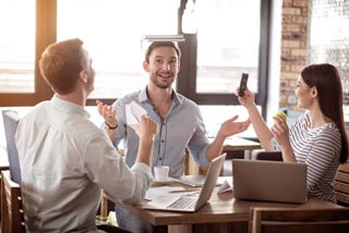 Three young professionals enthusiastically discussing while sitting at a table in a bright cafe with a laptop and coffee cups visible.