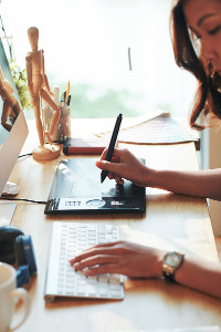 Woman using a graphics tablet and computer at a sunlit desk with a coffee cup and creative supplies