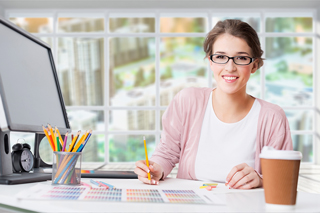 Young woman graphic designer smiling at the camera while working in a bright office, surrounded by colorful pencils, a computer monitor, and a coffee cup.