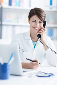 Medical admin assistant in lab coat talking on phone at desk with laptop and pen in hand