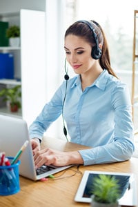 Woman in a collared shirt wearing a headset while typing on an open laptop at a wooden desk in a light-filled home office