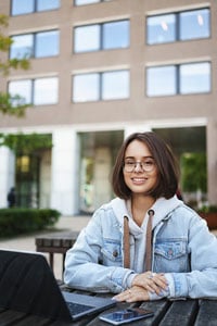 Young woman in glasses and denim jacket sitting at an outdoor table with a smartphone, smiling, with a modern building in the background.