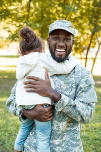 African American soldier in camouflage uniform holding a young child in a park.