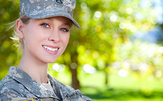 Smiling young woman outside in a camouflage military uniform with green trees and grass in the background