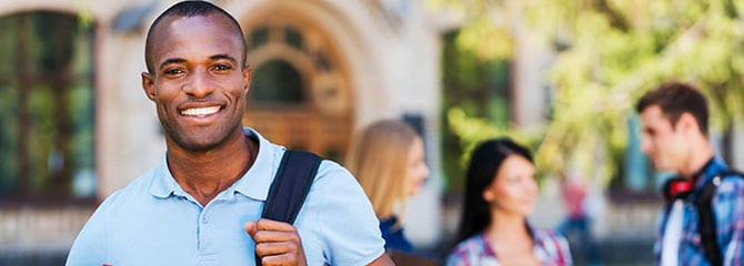 Smiling male college student holding a strap of his backpack and standing outside as three other students talk behind him
