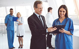 Hospital scene with two healthcare professionals, discussing a document on a clipboard, with other medical staff conversing in the background.