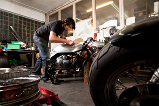 Man working on motorcycle in a garage surrounded by various auto parts and tools.