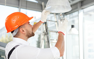 Electrician in orange hard hat installing a ceiling light fixture in a modern building