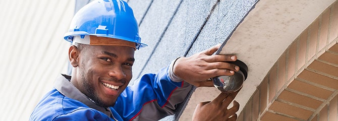 Smiling young man in a blue hard hat and work uniform inspecting a building's exterior light while standing on a ladder