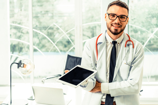 Doctor with beard wearing glasses, a white coat, and a stethoscope, holding a tablet in a bright office.