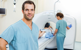 Radiology technician smiling in front of MRI machine with patient and colleague in background in hospital setting.