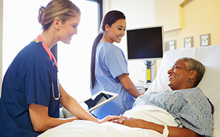 Two nurses attending to an elderly patient in a hospital bed, one holding a digital tablet and the other comforting the patient.
