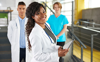 Confident nurse with stethoscope holding a clipboard in hospital corridor, accompanied by a diverse team of healthcare professionals in the background.