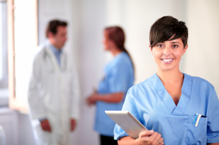 Nurse in blue scrubs holding a tablet with male and Doctors discussing in the background in a hospital corridor