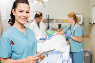 Nurse holding a tablet in a hospital room with colleagues treating a patient in the background.