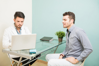 Two professional men in a meeting, one using a laptop and the other listening attentively, in an office with a glass table and light green wall.