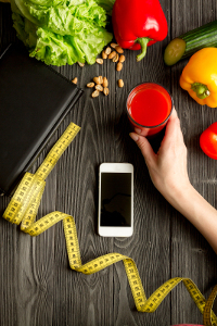 Overhead view of a woman's hand holding a glass of juice, surrounded by fresh vegetables, nuts, a measuring tape, smartphone, and notebook on a wooden table, symbolizing healthy eating and diet planning.