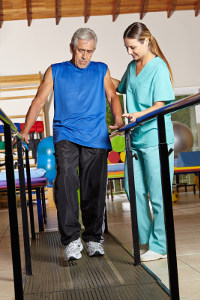 Senior man using walker for rehabilitation with assistance from female physical therapist in clinic gym.