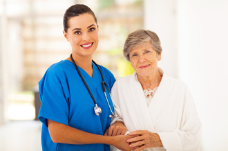 Nurse in blue scrubs with a stethoscope around her neck smiling and supporting an elderly woman in a white cardigan, both standing in a brightly lit corridor.