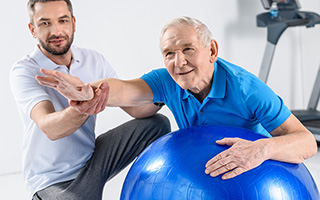 Elderly man with physical therapist doing exercises with a blue exercise ball in a gym setting