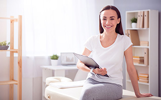 Physical therapist waiting for her patient while sitting on massage therapy bed in a bright clinical setting.