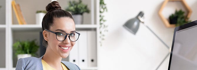 A smiling woman sitting at a computer, with shelves, plants, and a desk lamp in the background