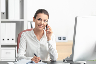 Young office administration professional talking on smartphone and writing notes at her desk in an office.