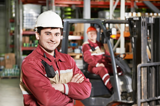Young male warehouse worker in red coveralls and white safety helmet smiling, with a colleague operating a forklift in the background.
