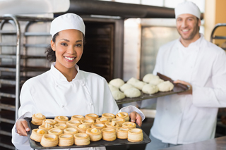 Two smiling bakers in white chef uniforms displaying freshly baked pastries in a bakery kitchen.