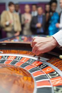 Close-up of a hand spinning a roulette wheel at a casino with blurred spectators in the background.