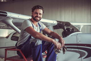 Smiling mechanic with a beard sitting on a step ladder next to a small aircraft in a hangar, holding a rag.