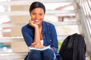 Young female student sitting on steps with a notebook, smiling at the camera, with a backpack beside her.