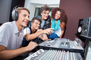 Four young adults smiling and interacting around an audio mixing console in a recording studio, with one wearing headphones.