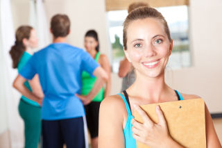 Fitness instructor holding clipboard with group of people talking in background at a gym.
