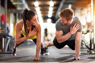 Two people doing push-ups in a gym, a woman in a yellow tank top and a man in a gray shirt, having a friendly competition or conversation.