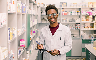 Pharmacy assistant in white coat holding clipboard in pharmacy with shelves of medication in background