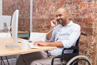 Man in a wheelchair working at a desk with a computer and talking on the phone in an office.