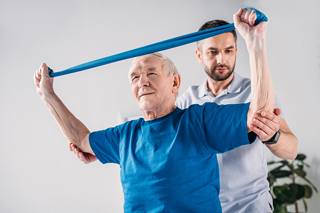 Elderly man exercising with resistance band assisted by physical therapist in clinical setting.