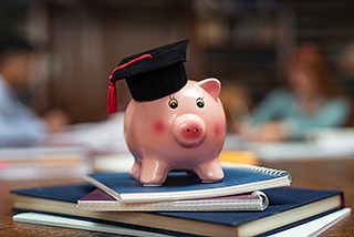 Graduation cap on piggy bank placed on top of stacked notebooks with blurred people in background indicating education savings or investment concept.