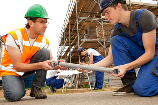 Two construction workers in safety gear assembling pipes on a construction site, with scaffolding in the background.