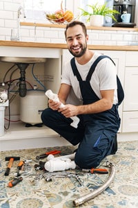 Smiling plumber in overalls holding a pipe fitting while sitting on the floor with various tools around him in a kitchen setting.