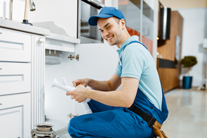 Smiling plumber in blue uniform and cap fixing sink and looking at camera in a modern kitchen