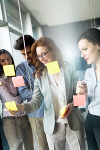 Group of four business professionals in discussion, using sticky notes on a glass wall in a bright office setting.