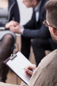 Close-up of a man in glasses holding a clipboard at a meeting with other professionals blurred in the background.