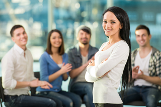 Young woman standing confidently  in front of a group of seated smiling colleagues in a bright office environment.