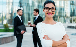 PR professional standing confidently in front of a glass building, with two men conversing in the background.
