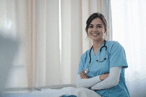 Smiling nurse wearing blue scrubs and a stethoscope, standing with arms crossed in a hospital room.