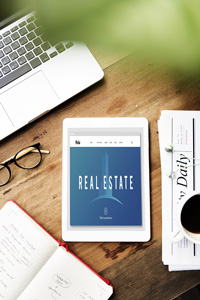 Wooden desk with laptop, tablet displaying "Real Estate" screen, glasses, coffee cup, and notebook