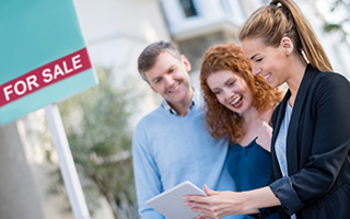 Real estate agent showing digital tablet to smiling couple near a 'For Sale' sign outside a house.