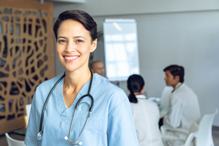 Registered nurse with stethoscope, wearing blue scrubs, standing in a hospital with colleagues in the background.